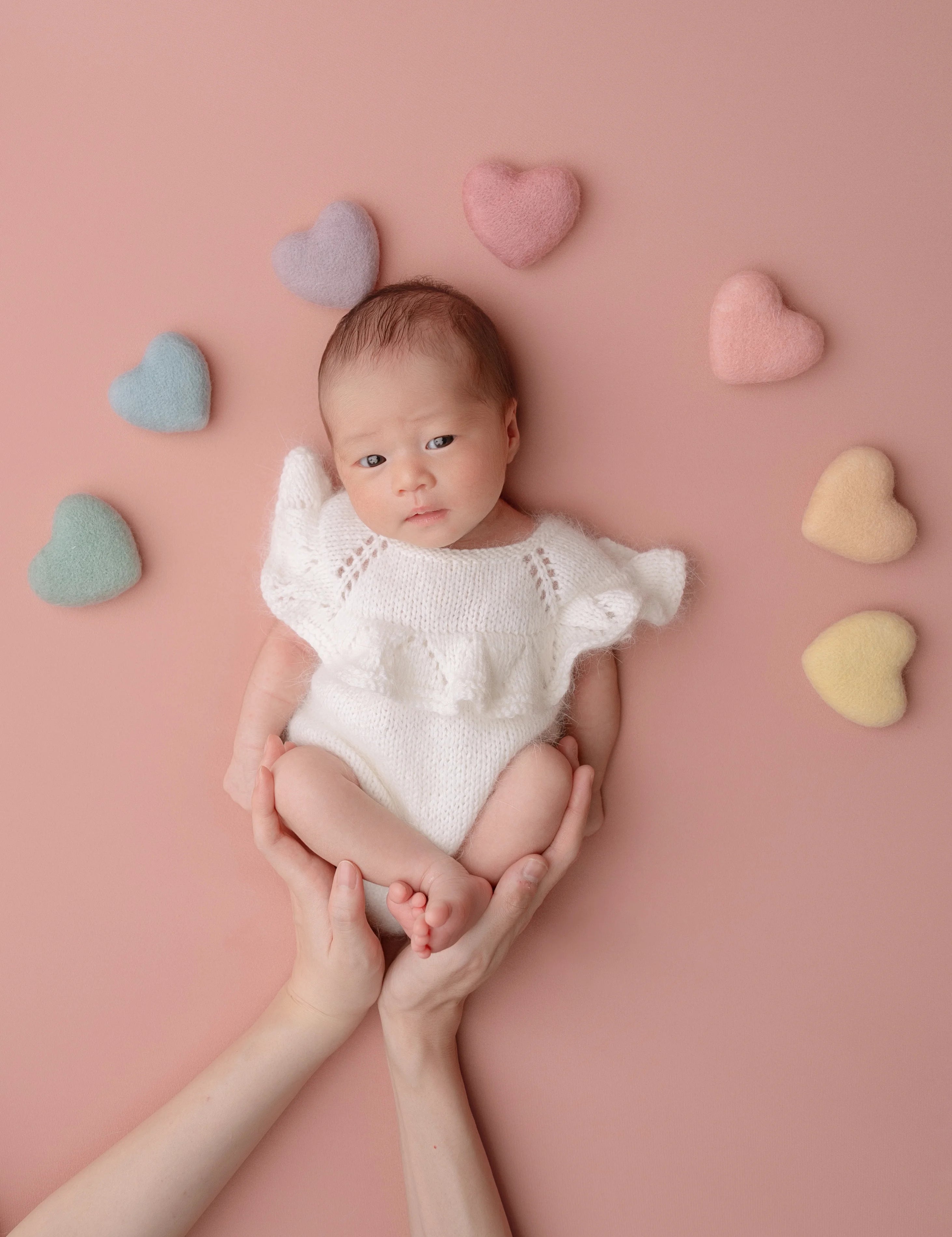 Newborn baby in a white outfit surrounded by colorful heart-shaped cushions on a pink background