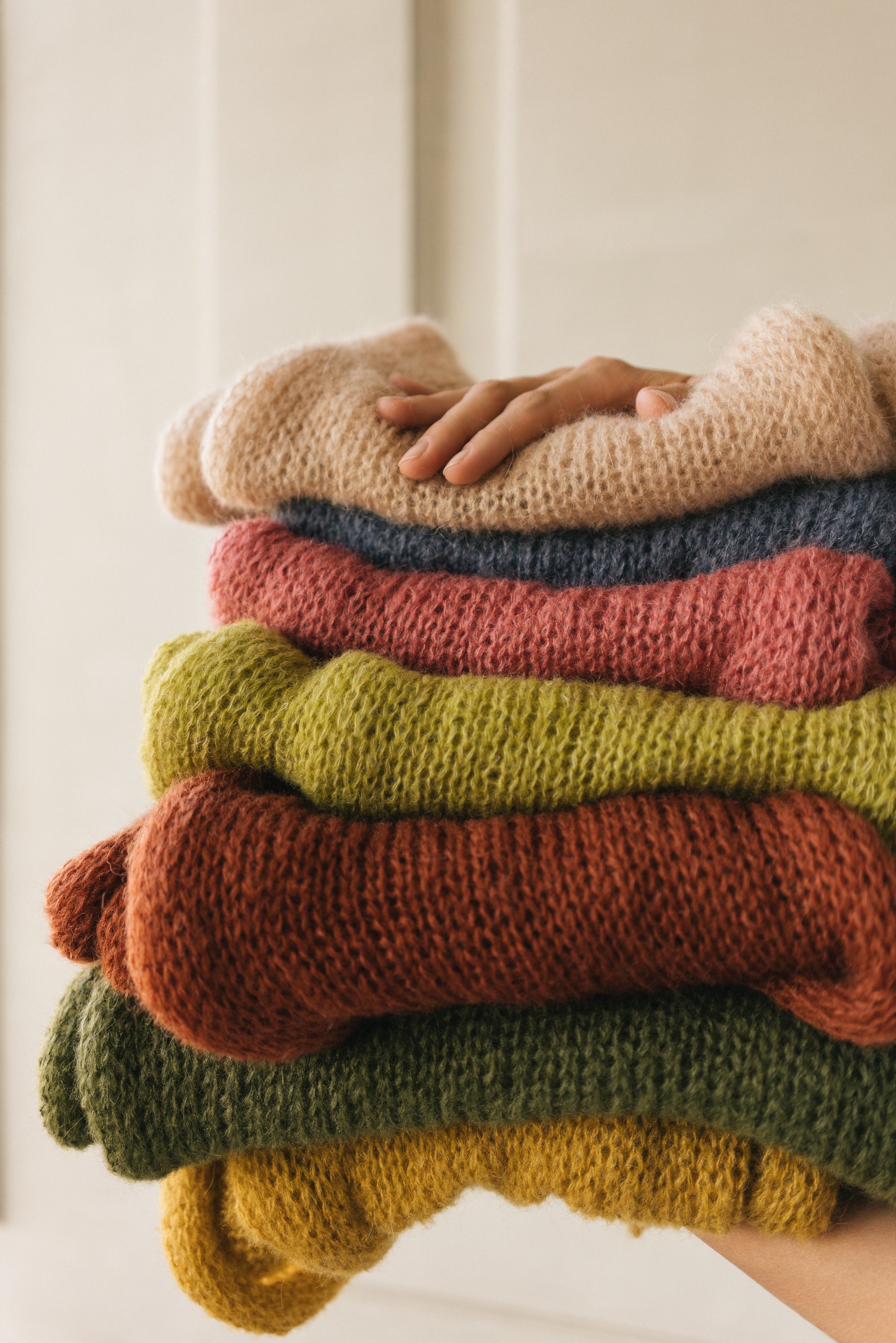 Stack of colorful sweaters with a hand on top against a neutral background