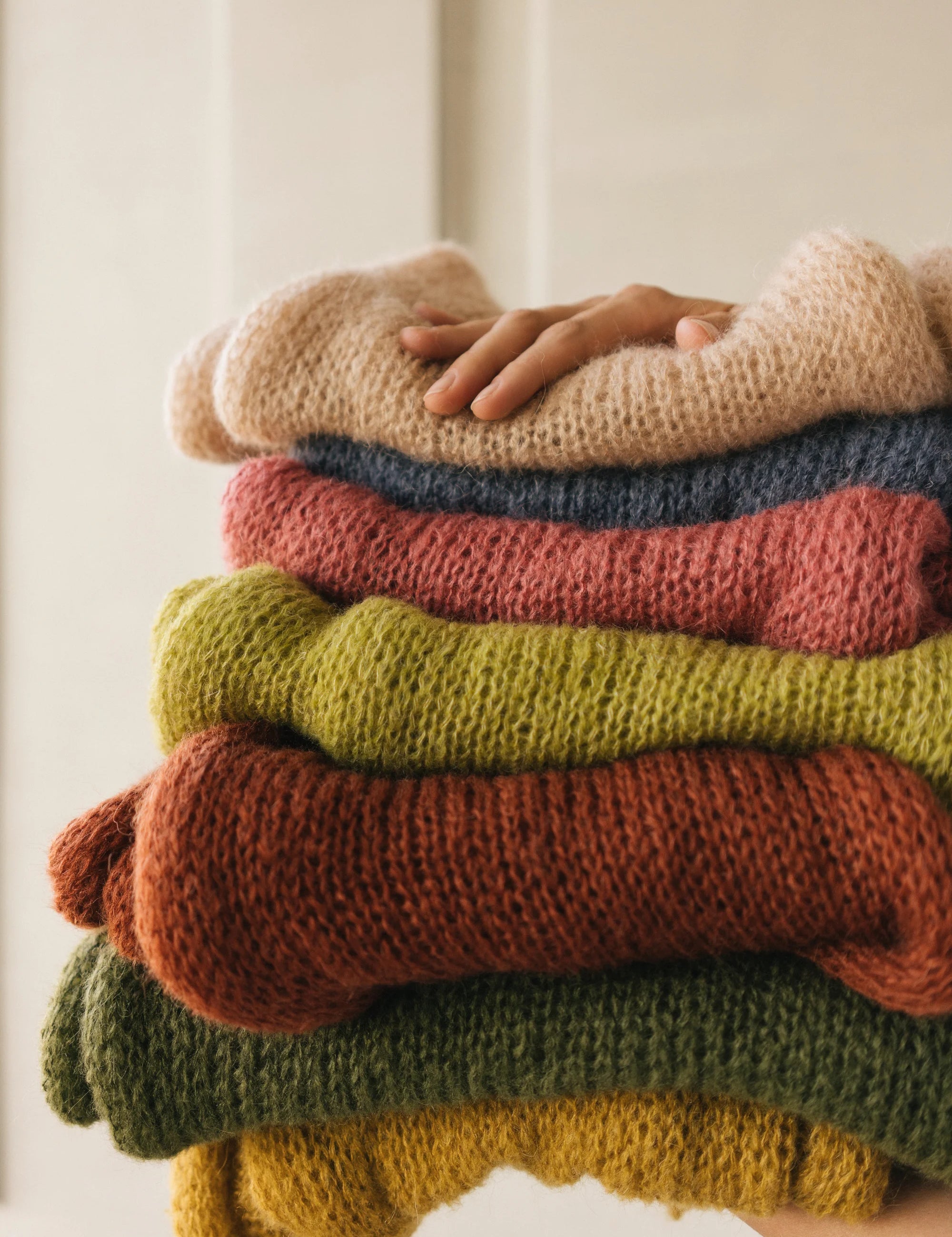 Stack of colorful sweaters with a hand on top against a neutral background