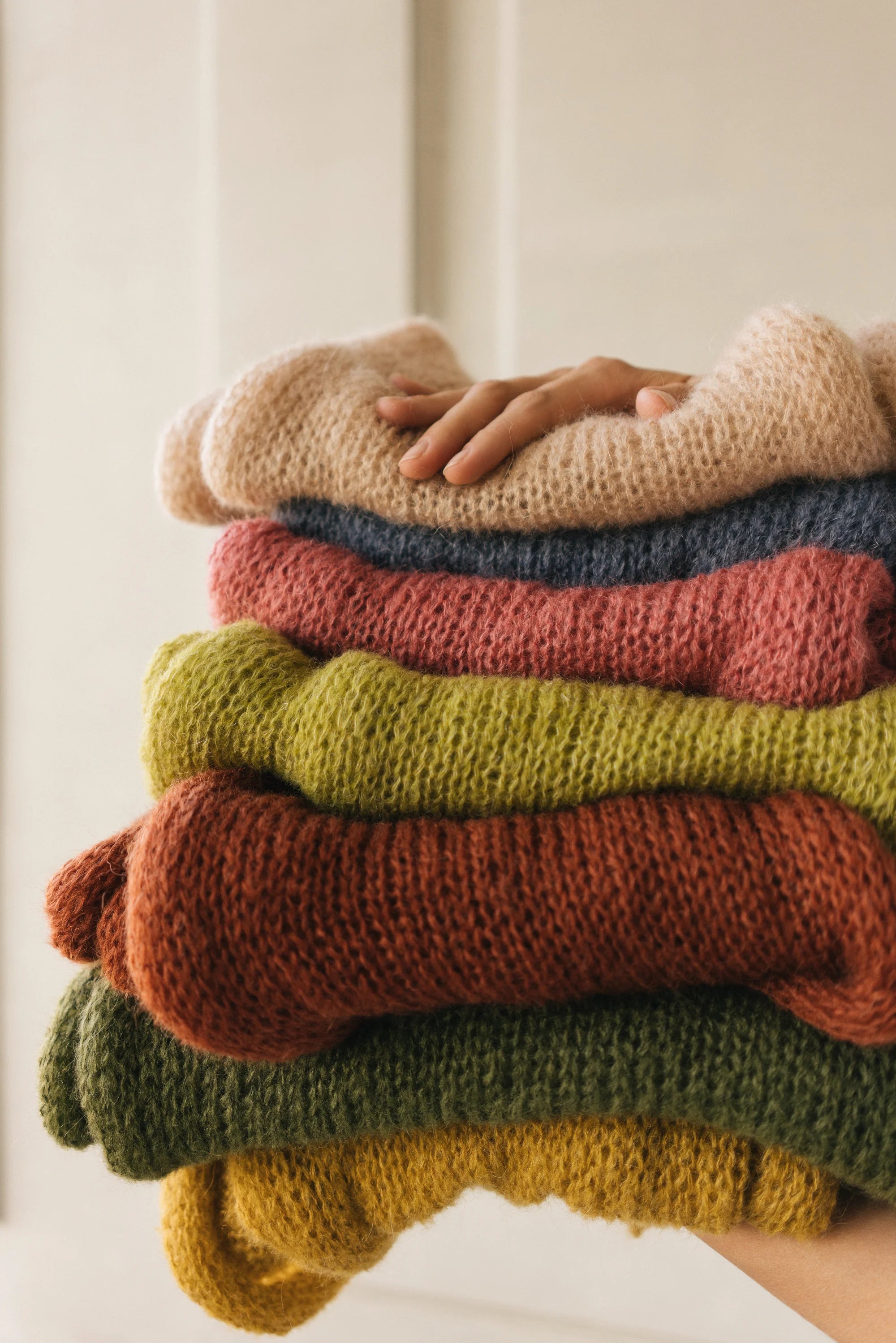 Stack of colorful sweaters with a hand on top against a neutral background