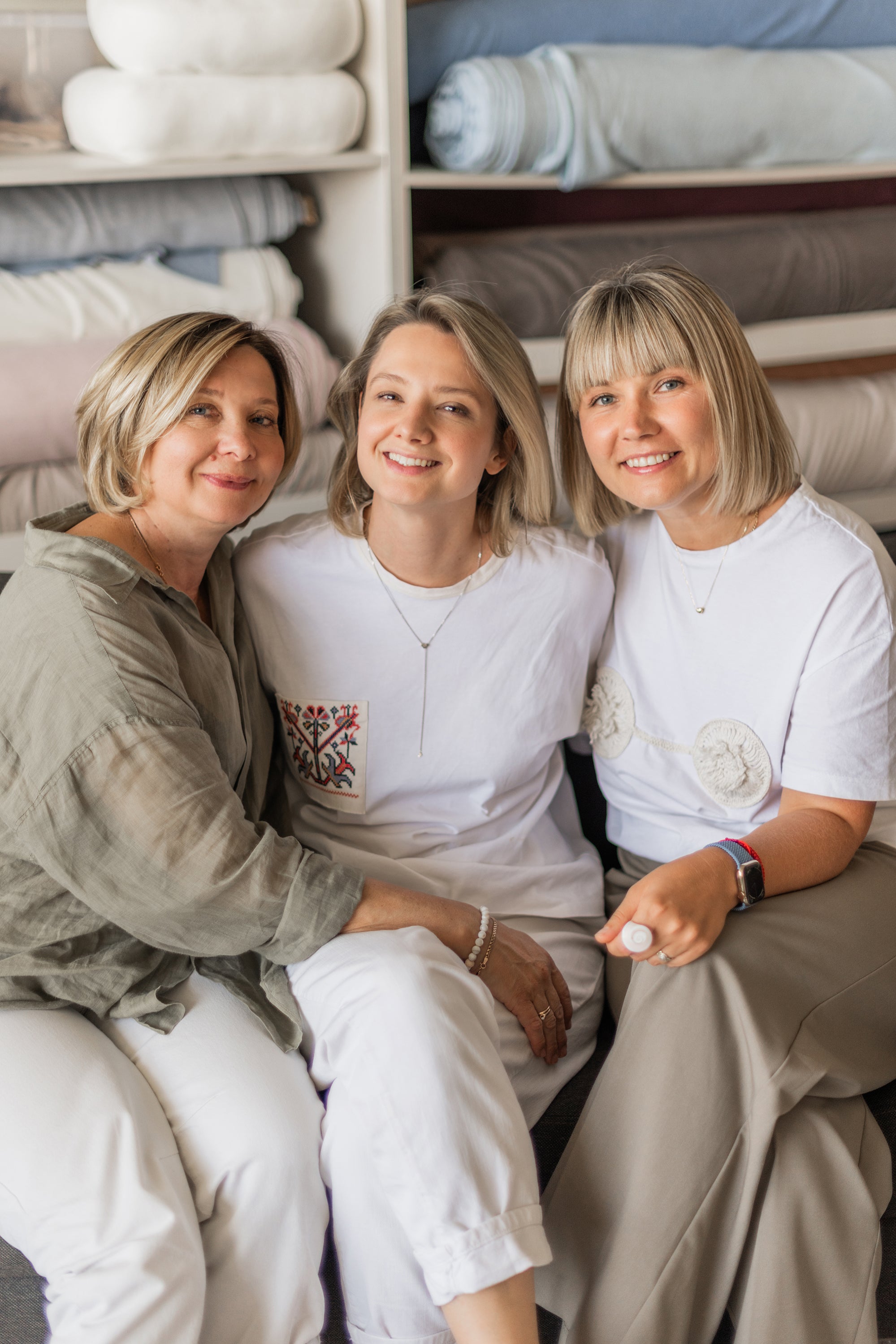 Three women sitting together in a fabric store with shelves of fabric behind them.