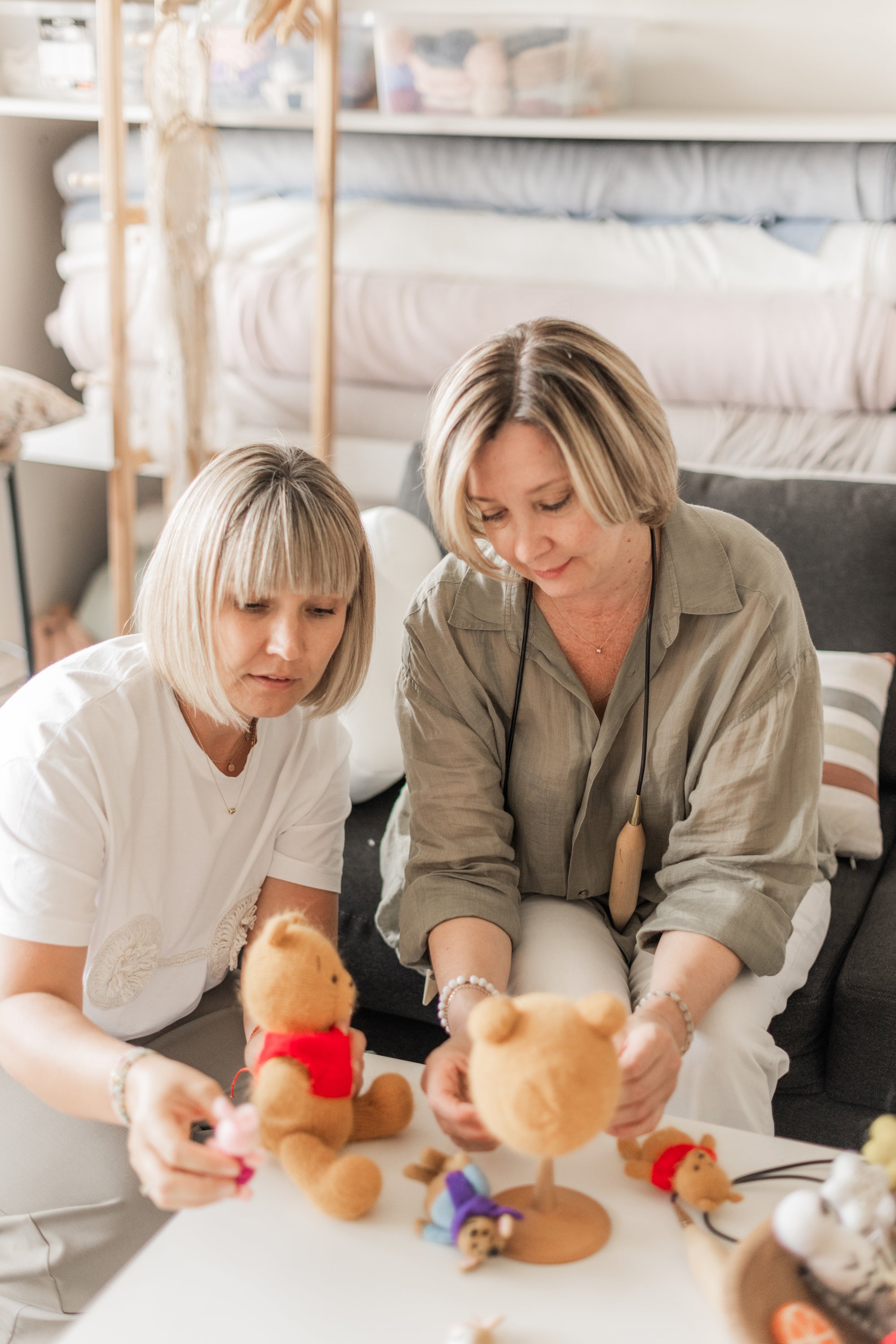 Two women sitting at a table with teddy bears and plush toys in a store setting.
