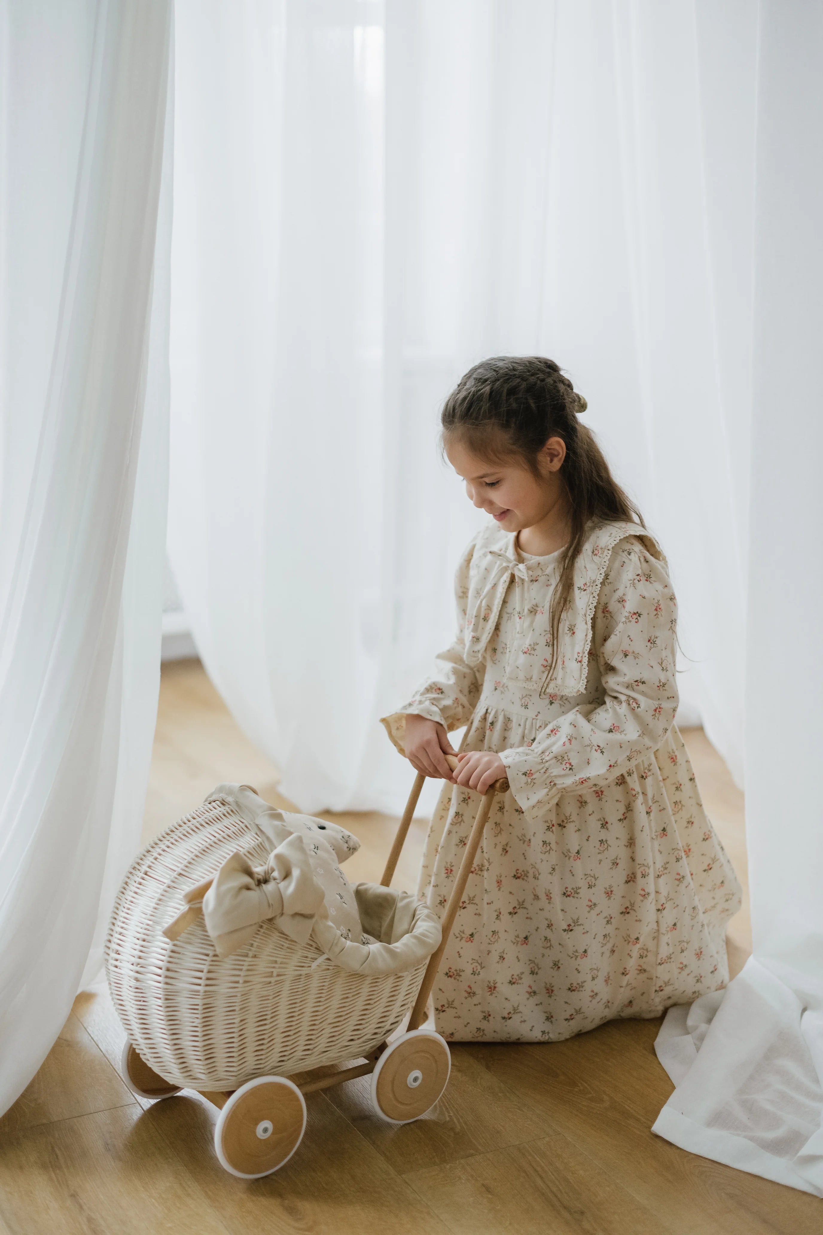 Young girl in a floral dress standing next to a wicker doll pram with a teddy bear inside, against a white curtain background.