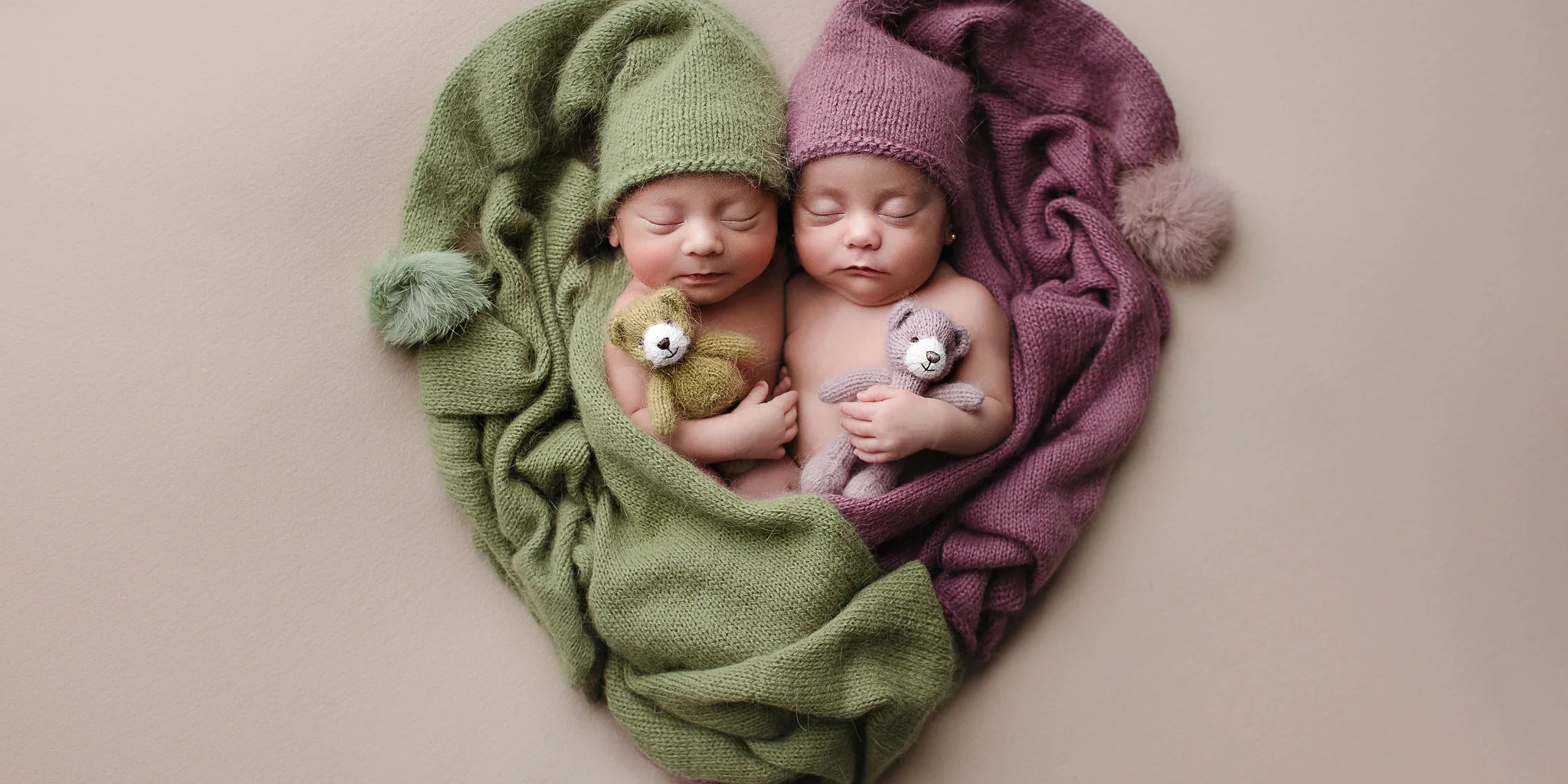 Two newborn babies wrapped in green and purple blankets with matching hats on a beige background.