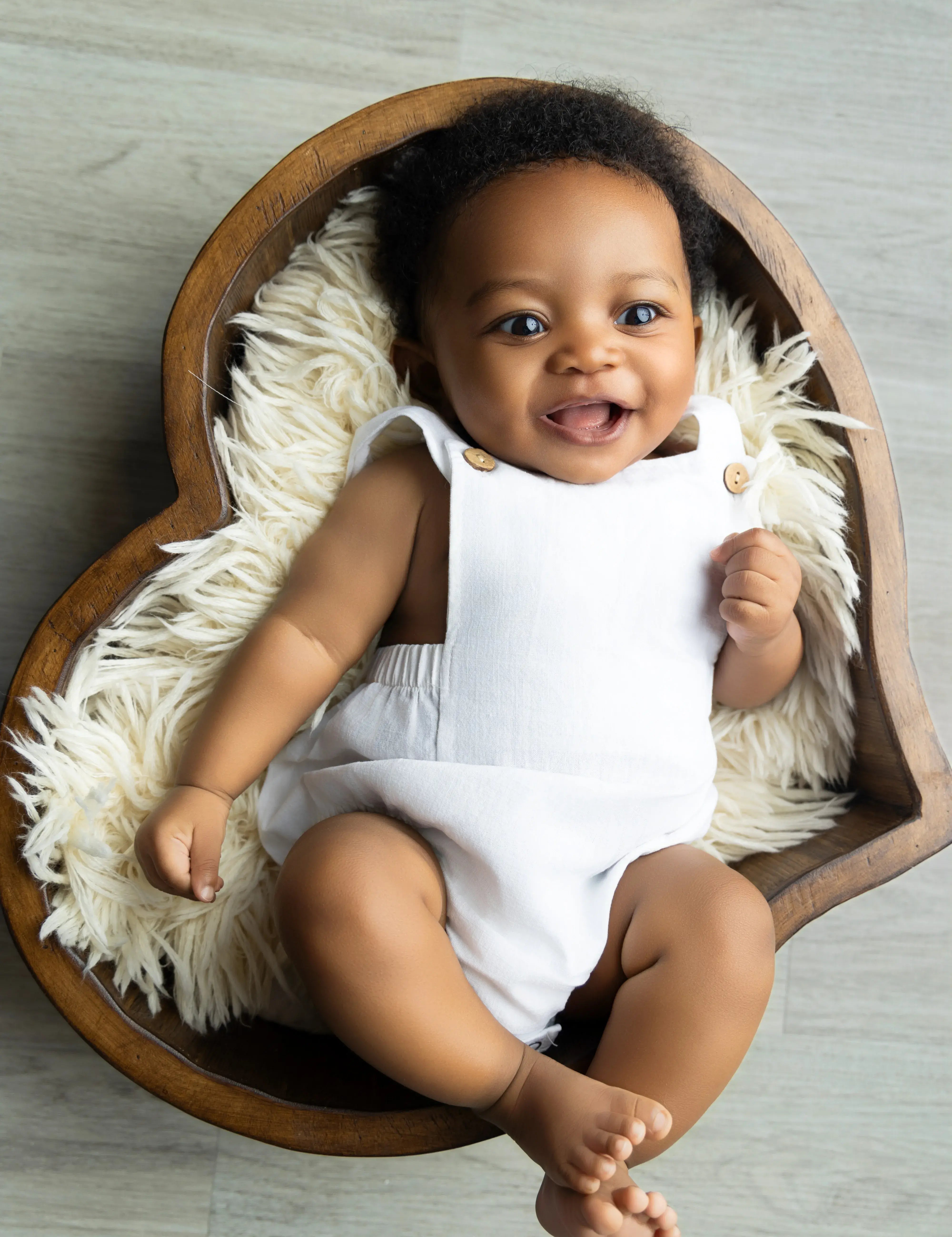 Baby in a white outfit sitting in a heart-shaped wooden bowl with a fluffy cushion on a light wooden floor.