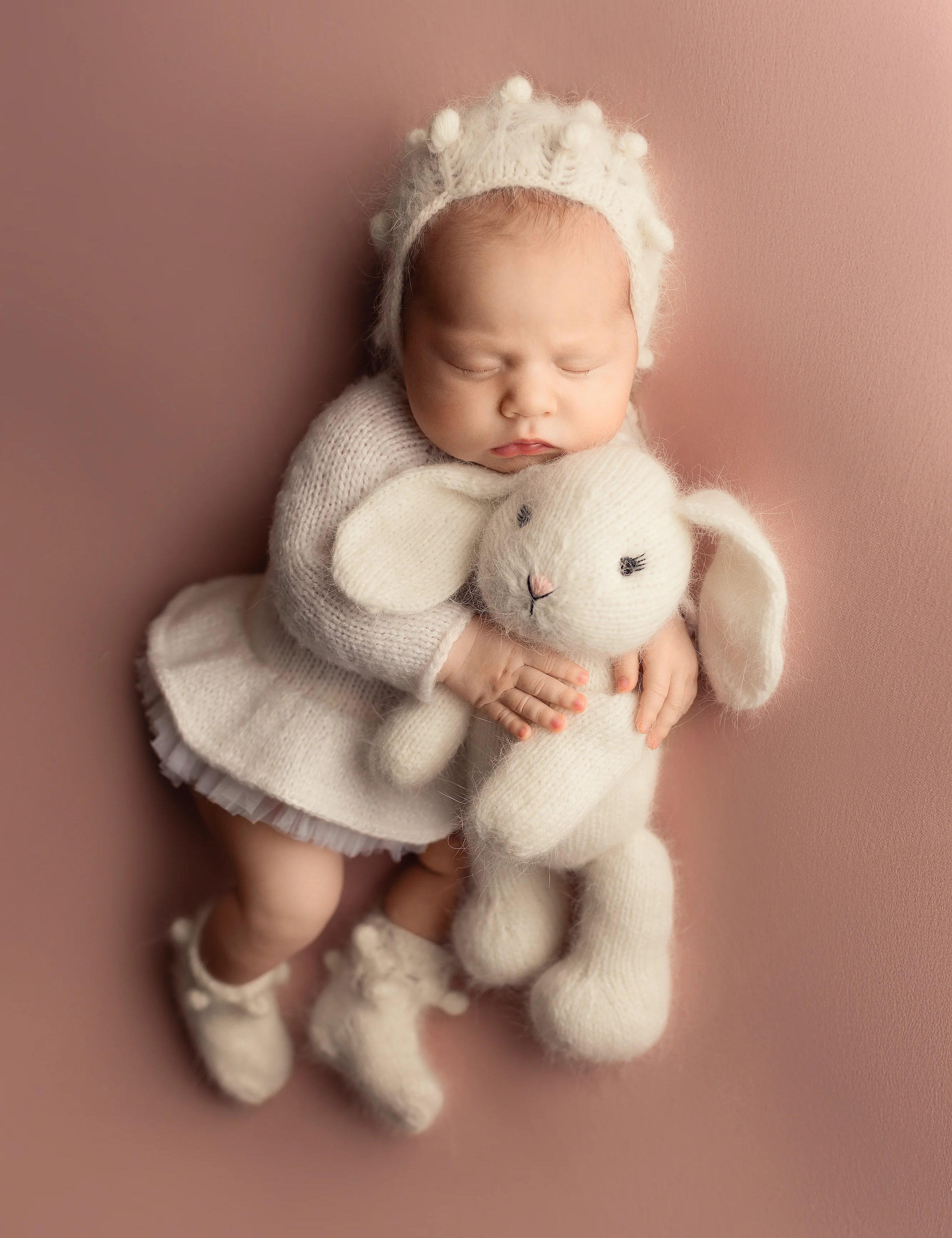 Newborn baby in a white outfit with a bunny hat and toy on a pink background