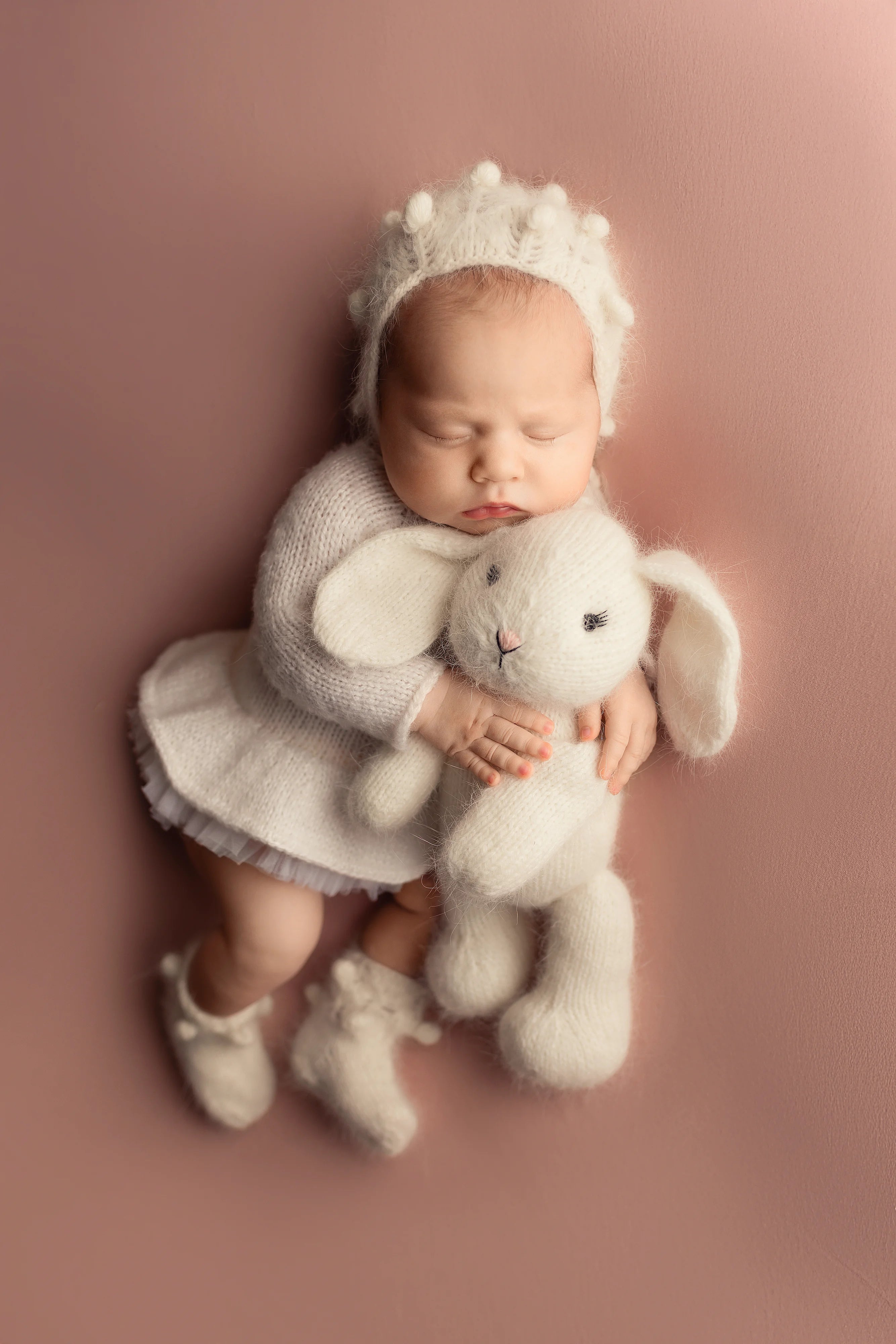 Newborn baby in a white outfit with a bunny hat and toy on a pink background