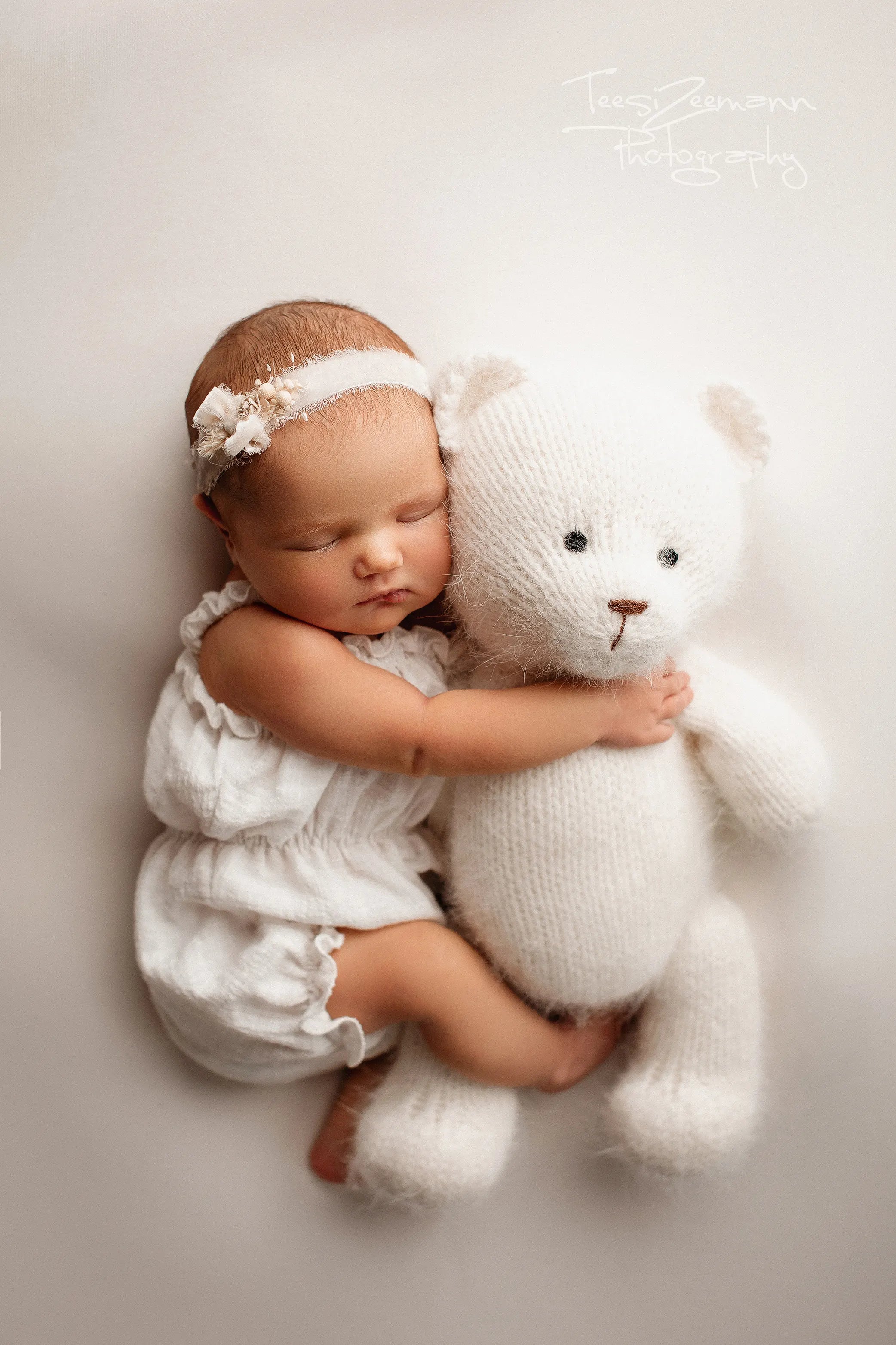 Newborn baby in a white outfit holding a white teddy bear on a light background