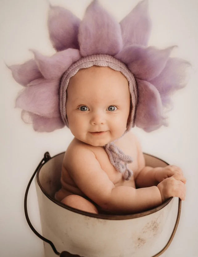 Baby wearing a purple flower hat sitting in a white bucket against a plain background