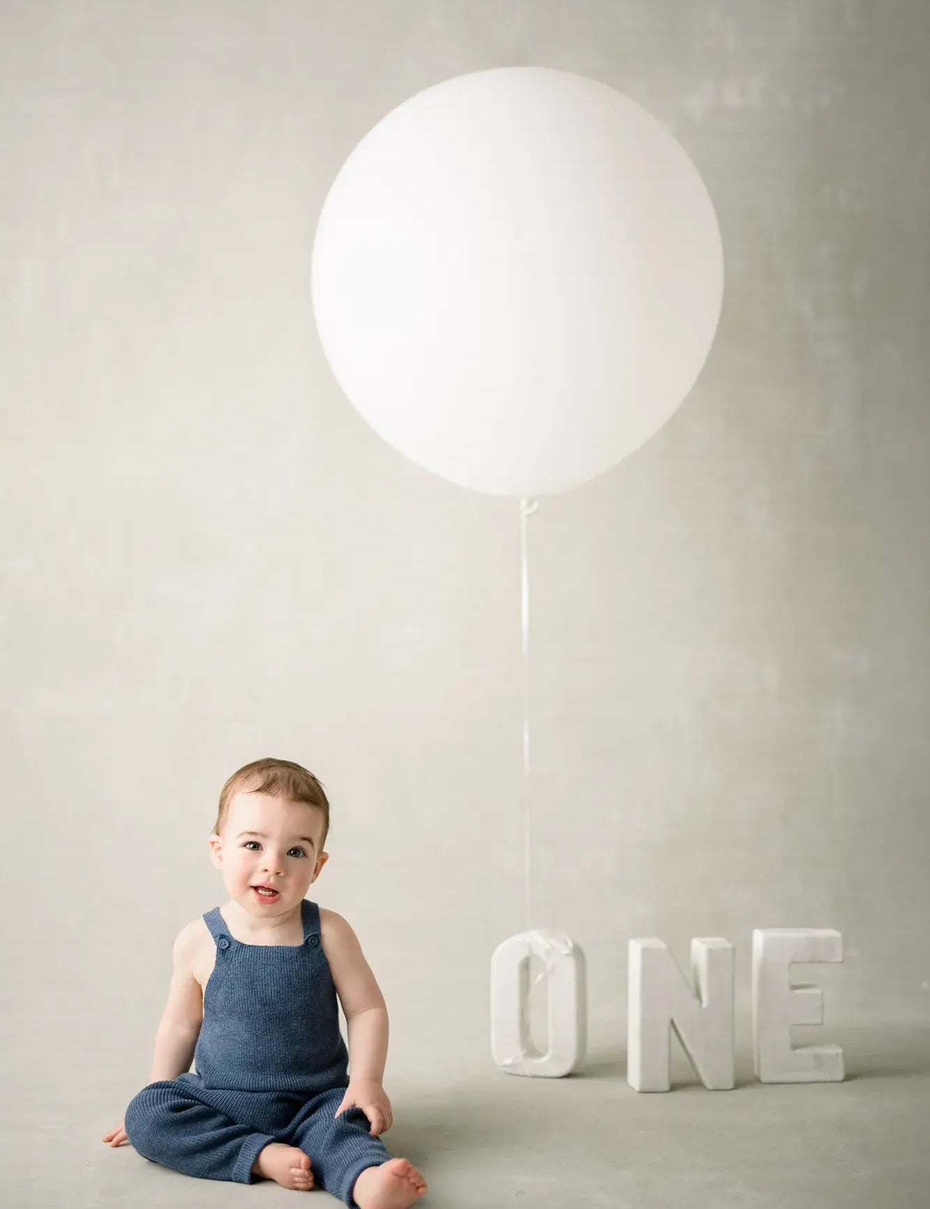 Child in a blue outfit sitting on the floor with a large white balloon and the word 'ONE' in the background.